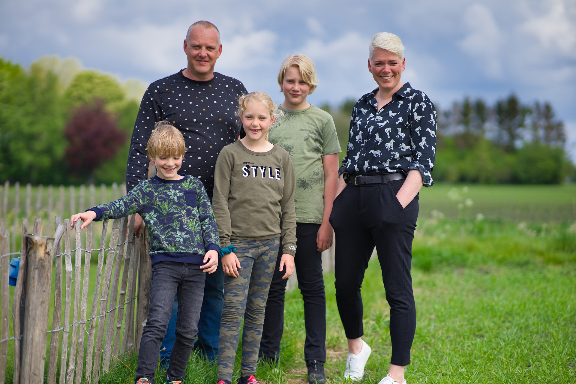 Familie steht am Zaun auf einer grünen Wiese, im Hintergrund Bäume und ein Campingplatzgelände.