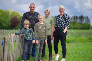 Familie steht am Zaun auf einer grünen Wiese, im Hintergrund Bäume und ein Campingplatzgelände.