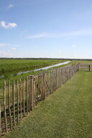 Grüne Wiese mit Holzzaun entlang eines schmalen Flusses, dahinter weite Felder unter blauem Himmel.