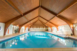 Innenpool mit blauem Wasser, Treppe und Holzdecke im Hallenbereich, Liegen und Stühle am Beckenrand.