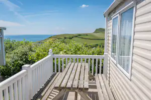 Wohnwagen-Terrasse mit Holzstufen und Geländer, Blick auf das Meer und grüne Hügel in der Ferne.