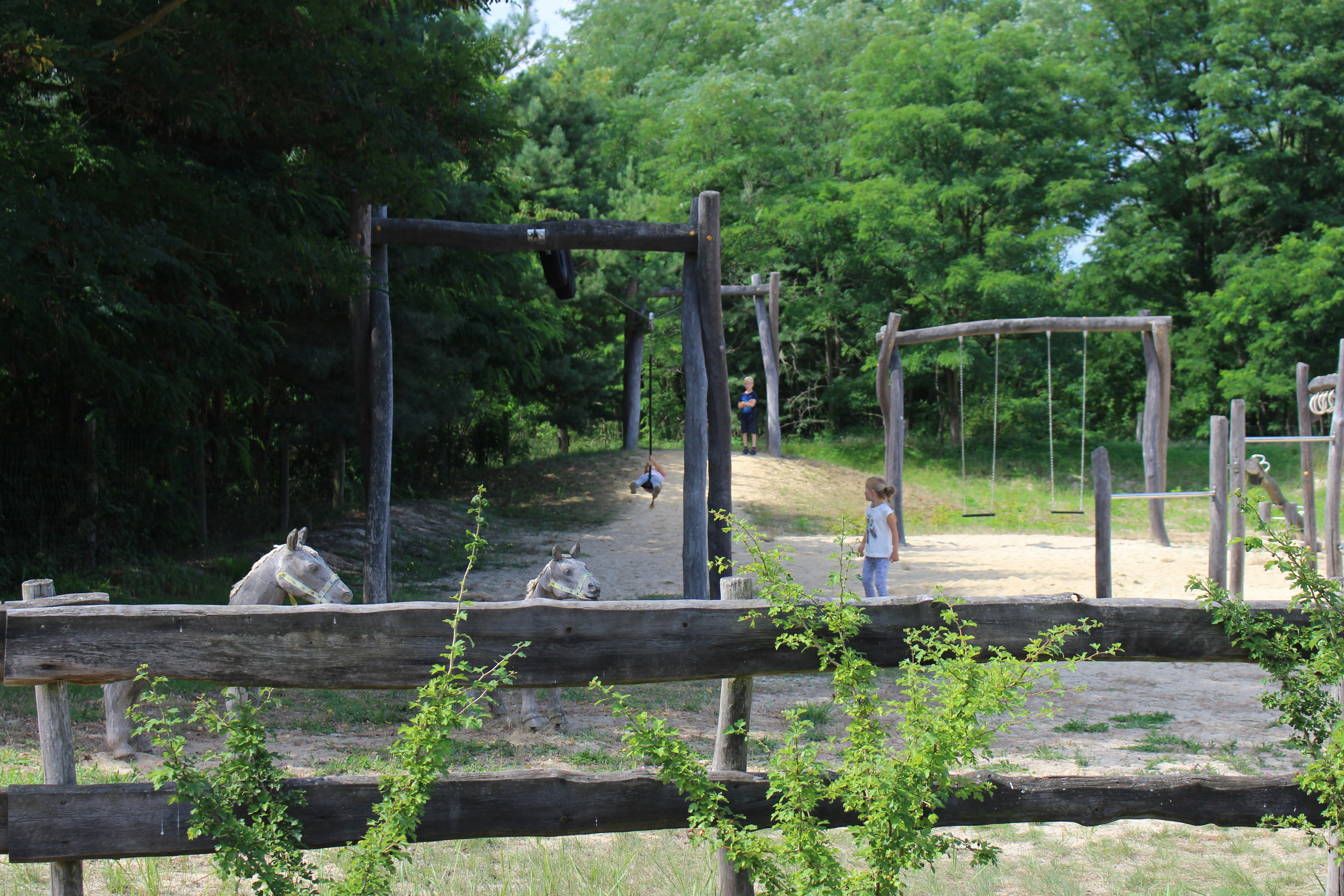 Spielplatz auf dem Campingplatz mit Holzschaukel, Sandboden und Bäumen im Hintergrund, daneben ein Weg und ein Zaun.