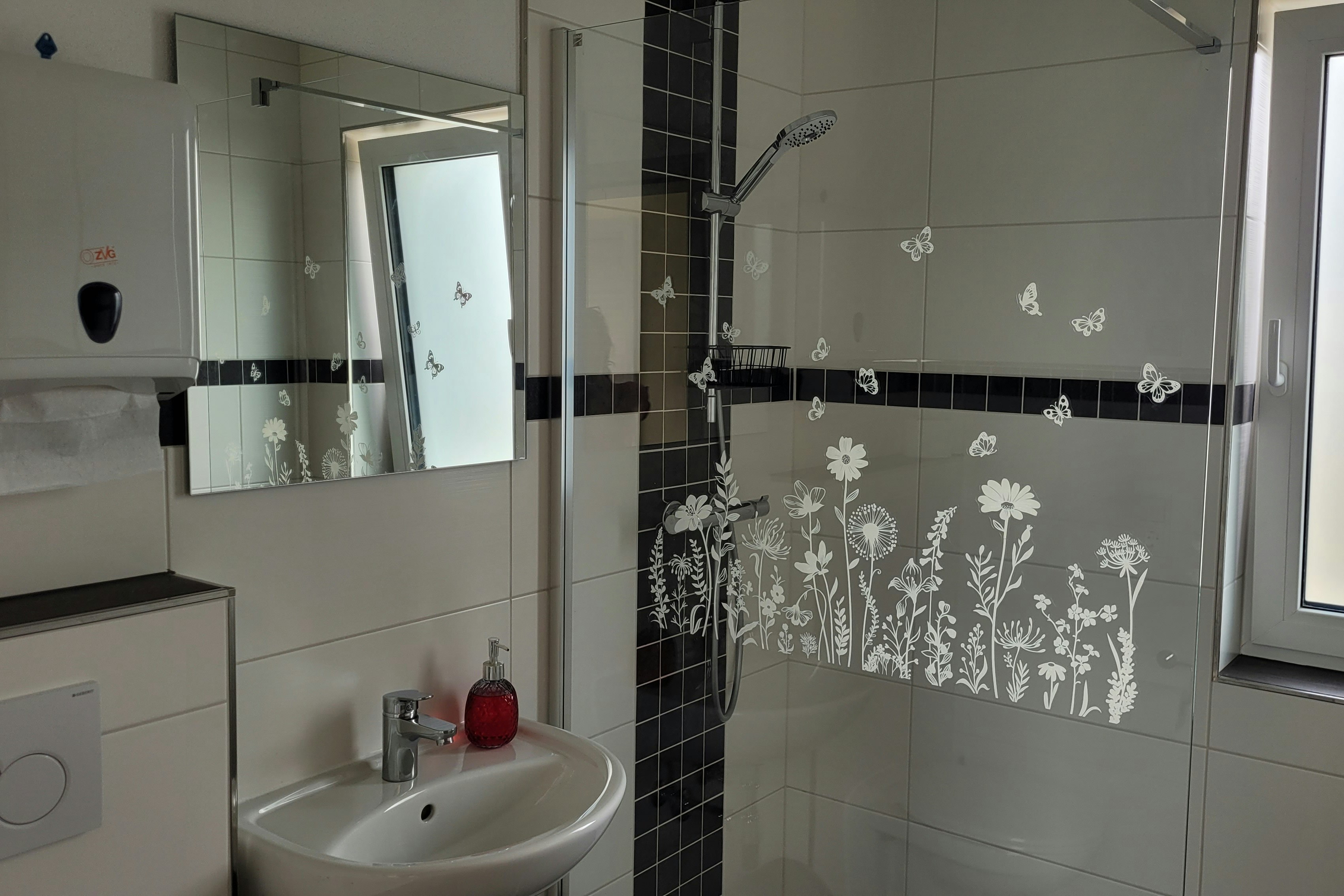 Modernes Badezimmer mit Waschbecken, Dusche mit Glaswand und Fliesen in Schwarz-Weiß, Fenster mit Tageslicht.