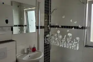 Modernes Badezimmer mit Waschbecken, Dusche mit Glaswand und Fliesen in Schwarz-Weiß, Fenster mit Tageslicht.