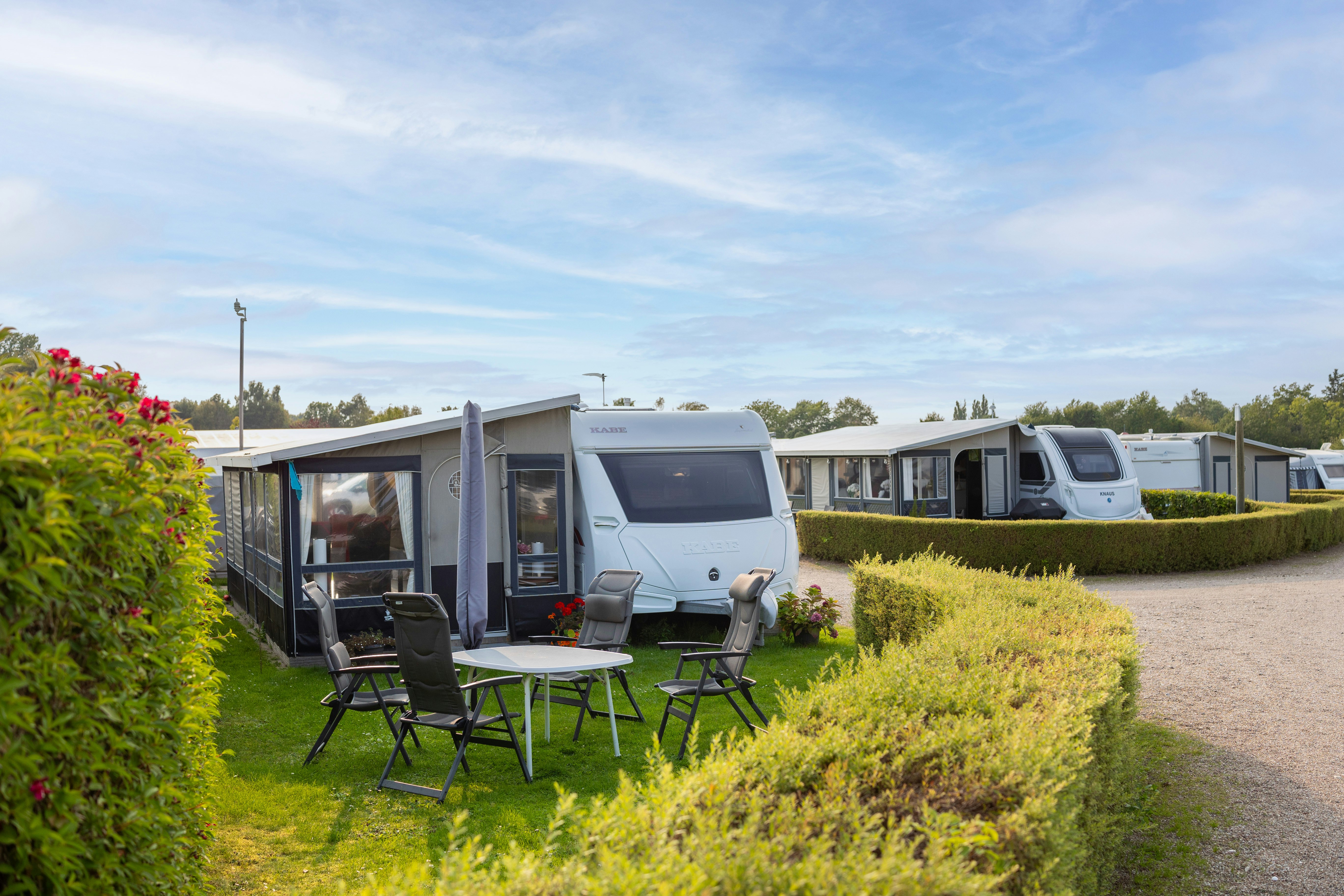 Wohnwagen auf einem Campingplatz mit Terrasse und Gartenmöbeln, Hecken und Kiesweg; im Hintergrund weitere Wohnmobile und Bäume.