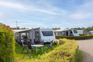 Wohnwagen auf einem Campingplatz mit Terrasse und Gartenmöbeln, Hecken und Kiesweg; im Hintergrund weitere Wohnmobile und Bäume.