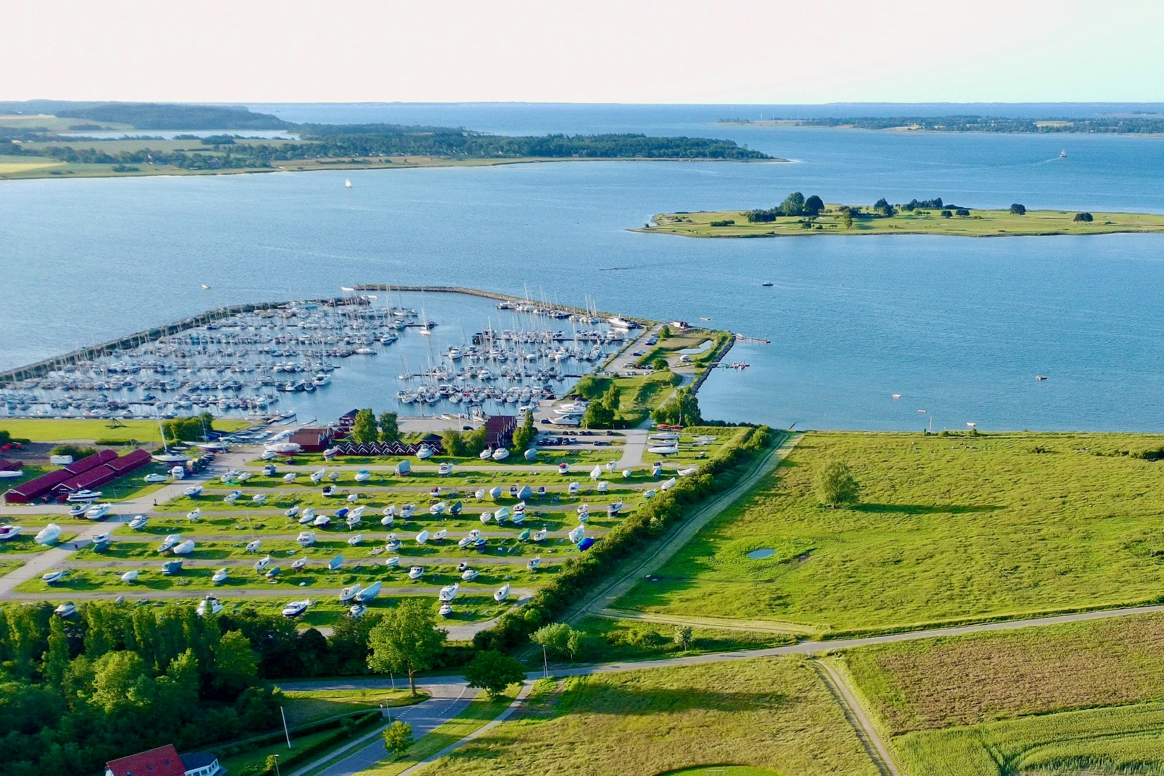 Luftaufnahme einer Küstenlandschaft mit Bucht, grünen Wiesen, Wegen und einem kleinen Ort am Wasser.