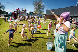 Kinder und Familien spielen auf einer Campingwiese, im Hintergrund ein Spielplatz mit bunten Hüpf- und Spielgeräten.