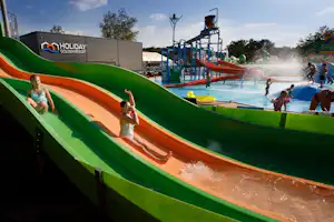Grüne Wasserrutsche auf dem Campingplatz, Kinder rutschen ins Becken, im Hintergrund Gebäude und Spielbereich.