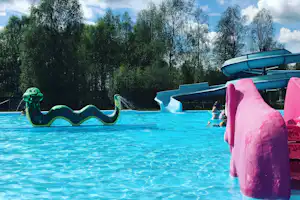 Blauer Pool mit Rutsche und aufblasbaren Figuren, im Hintergrund Bäume und ein Zaun am Campingplatz.