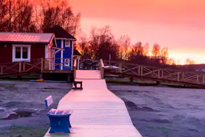 Holzterrasse mit Weg zum Campinghaus, Geländer und Sitzbank, im Hintergrund weitere Häuser und Bäume bei Sonnenuntergang.