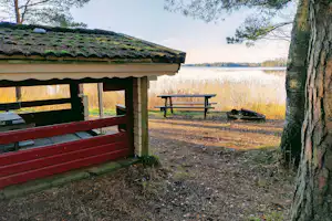 Campinghütte mit roter Holzveranda, Bank und Tisch am Weg, im Hintergrund ein See und Schilf.