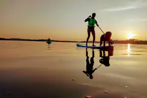 Stand-up-Paddler auf dem See bei Sonnenuntergang, Spiegelung im Wasser und ein weiterer Paddler im Hintergrund.