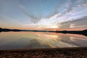 Sonnenuntergang am See mit Spiegelung im Wasser, Sandstrand im Vordergrund und ruhige Uferlinie in der Ferne.