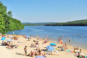 Badestrand am See mit vielen Familien, Sonnenschirmen und Liegestühlen, im Hintergrund Wald und Hügel.