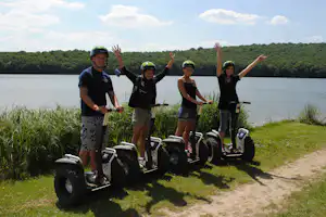 Gruppe mit Helmen auf Segways am Seeufer, im Gras und auf dem Weg, im Hintergrund Wald und Wasser.
