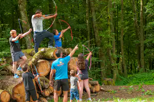 Familie beim Bogenschießen im Wald, Kinder und Erwachsene mit bunten Bögen vor Baumstämmen und grünen Bäumen.