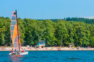 Windsurfsegel am See vor einem Campingstrand mit vielen Badegästen, Bäumen und einem Gebäude im Hintergrund.