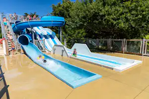 Blaue Wasserrutsche im Außenbereich mit Kindern auf der Treppe, Wasser läuft, Holzgeländer und Bäume im Hintergrund.