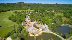 Luftaufnahme eines Campingplatzes am See mit Pool, Tennisplatz, Gebäuden, Wegen und Wohnwagen inmitten grüner Hügel.