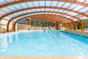 Beheizter Campingpool unter Holzbogen und Glasdach, mit Liegestühlen und Sitzbereich am Beckenrand.