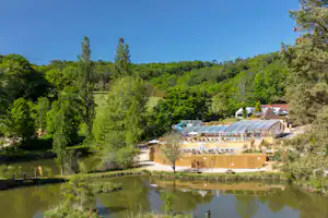 Campingplatz am See mit Holzgebäude und Terrasse, umgeben von Bäumen und Grünflächen, im Hintergrund ein weiteres Gebäude.