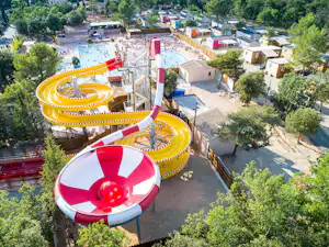 Blick auf gelbe und rote Wasserrutschen, einen großen Pool und Wohnhäuser im Campingbereich, umgeben von Bäumen.