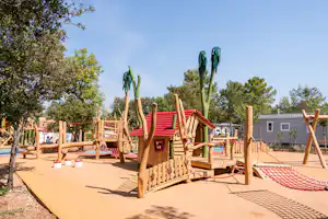 Holz-Spielhaus mit rotem Dach und Kletterelementen auf dem Spielplatz, umgeben von Bäumen und einem Weg.