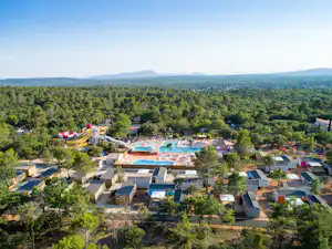 Blick auf Campinganlage mit Schwimmbecken, Wasserrutsche und vielen Bungalows, umgeben von Bäumen und Hügeln in der Ferne.
