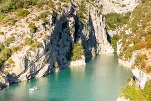 Türkisfarbenes Wasser zwischen steilen Felswänden, bewachsen mit Büschen und Bäumen; im Vordergrund kleine Boote.