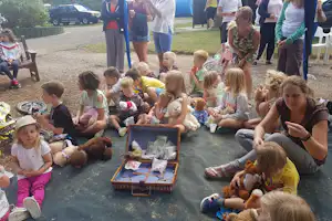 Kinder sitzen auf dem Campingplatz auf einer Decke und spielen mit Teddybären, während Erwachsene daneben stehen.