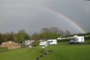 Wohnmobile auf einer Campingwiese, im Hintergrund Hecken und Bäume, darüber ein Regenbogen am Himmel.