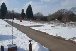 Campingweg im Winter mit Schnee, Hecken und Bäumen, roter Telefonzelle und Absperrpfosten am Rand.