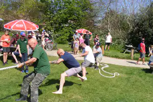 Campinggruppe spielt Tauziehen auf einer Wiese, im Hintergrund rote Sonnenschirme, Fahrräder und Bänke am Weg.