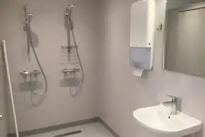 Dusche mit Brausekopf und Halterung, daneben Waschbecken und Wandarmaturen im hellen Sanitärraum.