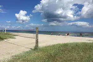 Strand mit Volleyballnetz am Sand, im Hintergrund Meer und Menschen am Ufer unter bewölktem Himmel.
