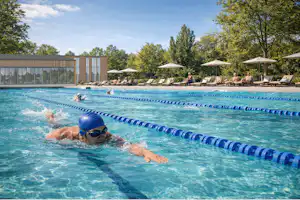 Schwimmer im blauen Badeanzug im Schwimmbecken mit Bahnen, im Hintergrund Liegen und Sonnenschirme am Campingplatz.