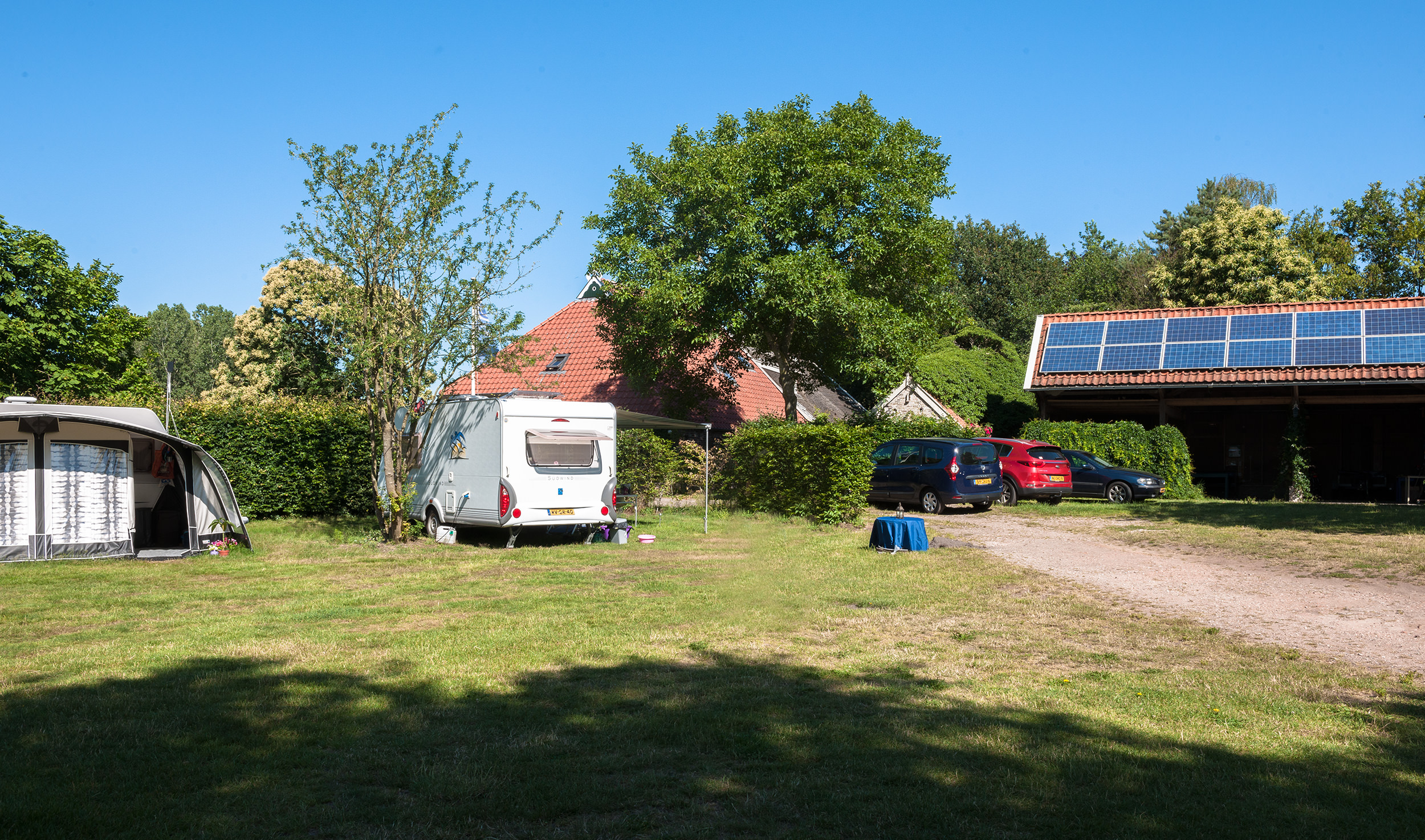 Stellplatz mit Wohnwagen und Zelt, Rasenfläche, Hecke, Sonnensegeln auf dem Dach und geparkten Autos am Weg.
