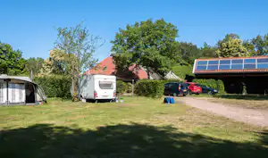 Stellplatz mit Wohnwagen und Zelt, Rasenfläche, Hecke, Sonnensegeln auf dem Dach und geparkten Autos am Weg.