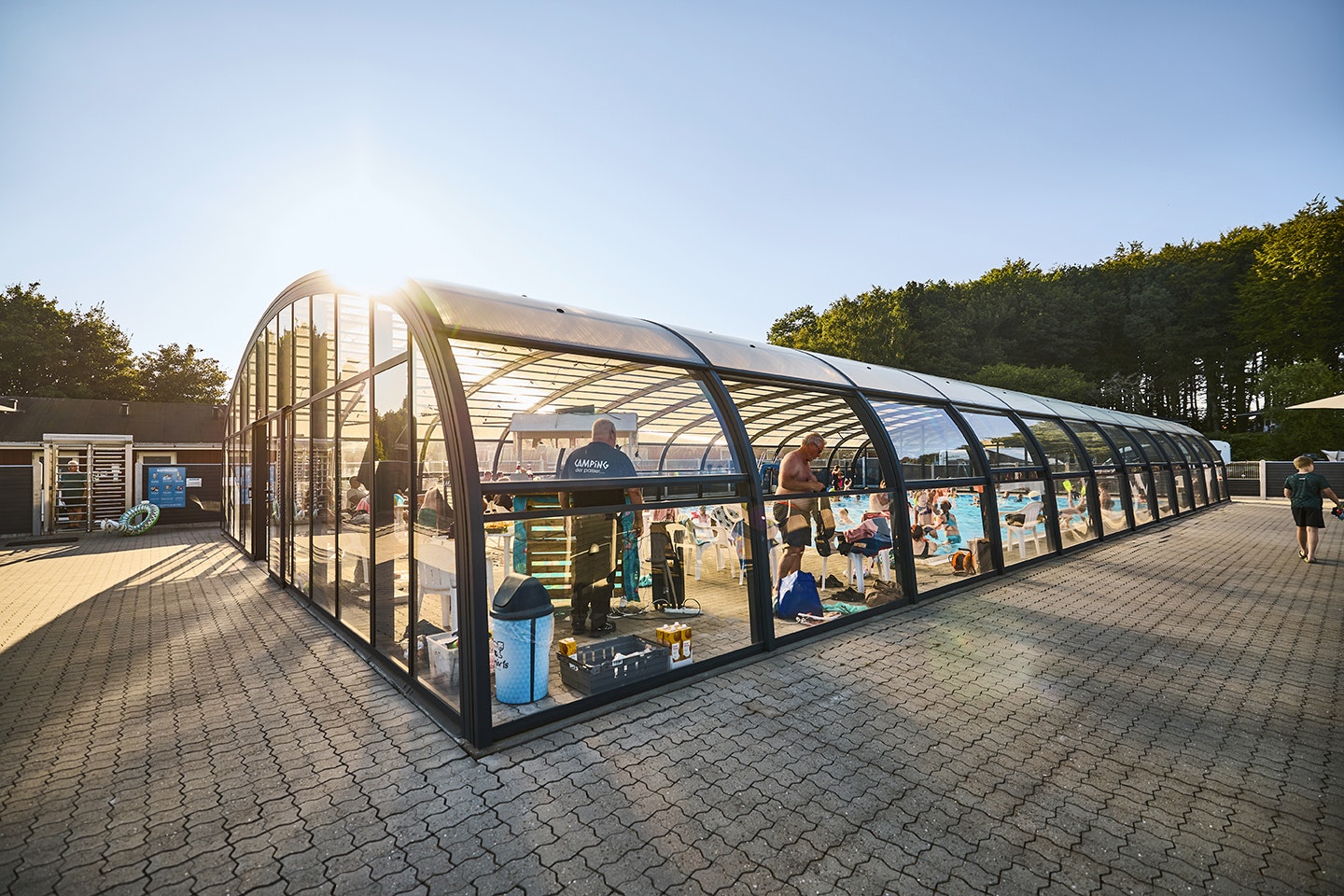 Überdachtes Schwimmbecken mit Glaswänden, Sitzgelegenheiten und Tischen auf dem gepflasterten Campingplatz, im Hintergrund Bäume.