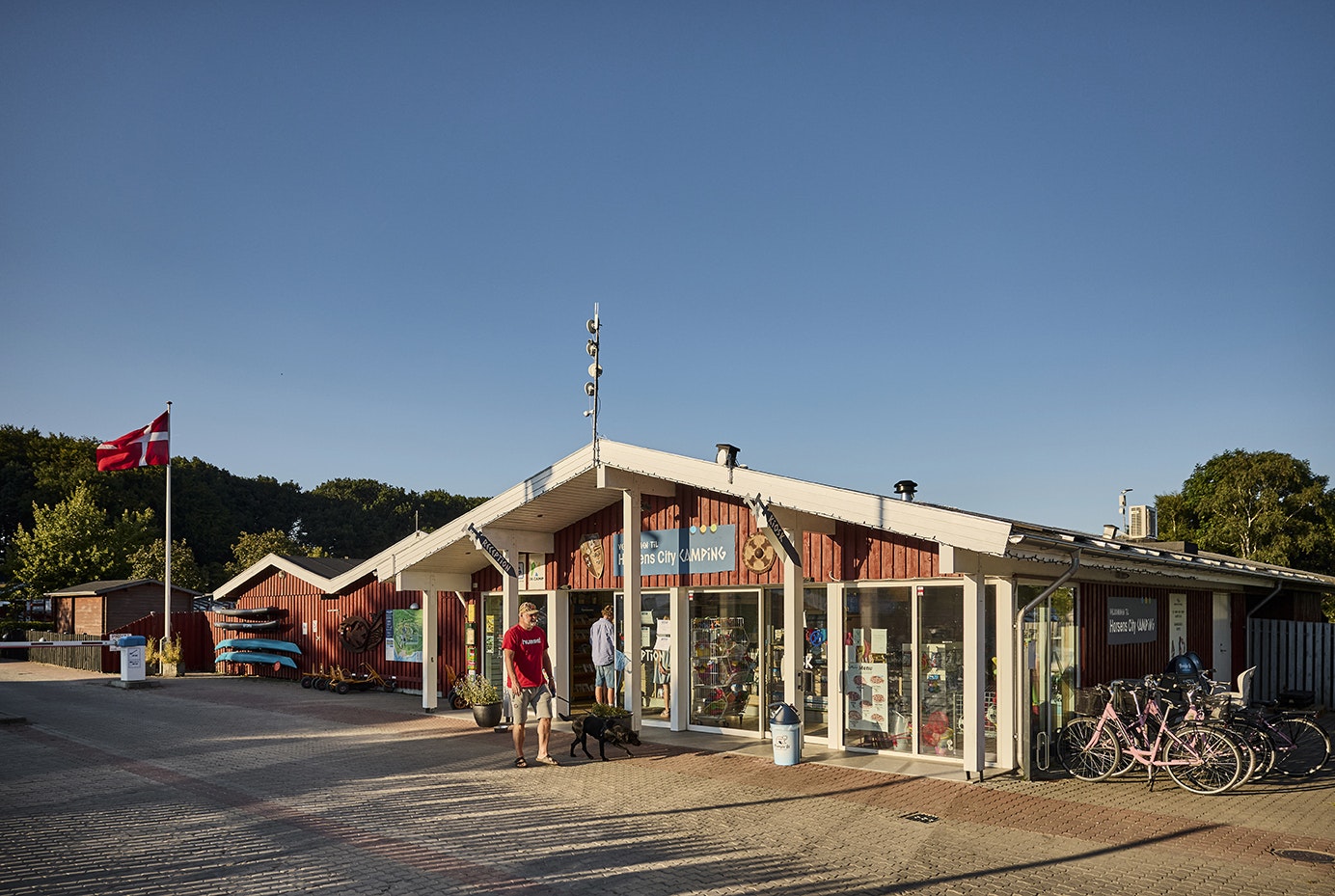 Eingang eines Campingplatzes mit Gebäude, Glasveranda und Sitzgelegenheiten, davor Pflasterweg und Fahrräder.