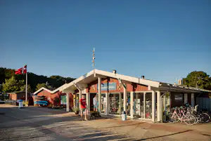 Eingang eines Campingplatzes mit Gebäude, Glasveranda und Sitzgelegenheiten, davor Pflasterweg und Fahrräder.