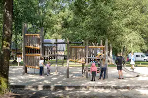 Holz-Spielplatz mit Rutsche und Klettergerüst auf Sand, Kinder spielen im Grünen neben Wohnwagen und Bäumen.
