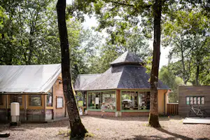Holzbar am Campingplatz unter Bäumen, daneben Zelt mit Planenwand und Kiesweg.
