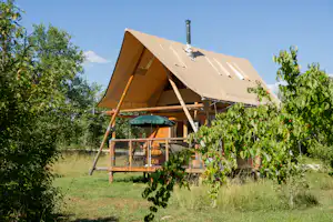 Campingzelt mit Veranda und Holzgestell im Grünen, umgeben von Büschen und Bäumen vor klarem Himmel.