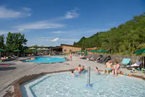 Großer Camping-Pool mit Wasserfontäne, Liegen und Sonnenschirmen, im Hintergrund Gebäude und Bäume.