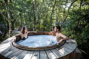 Whirlpool auf einer Holzterrasse im Grünen, zwei Personen im Wasser, umgeben von Bäumen und Gartenblick.
