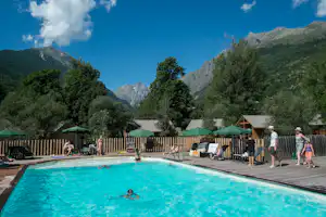 Camping-Pool mit türkisfarbenem Wasser, Liegen und Zaun, im Hintergrund Berge und Bäume unter blauem Himmel.