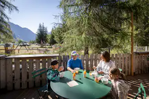 Familie sitzt an einem grünen Tisch auf der Holzterrasse, im Hintergrund Bäume, Campingplatz und Berge.