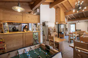 Camping-Bar mit Holzdecke, Theke, Getränken und Kicker-Tisch; daneben Holztische und Stühle im Innenraum.